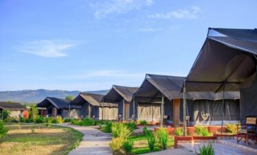 A row of budget safari tents with black canvas roofs and private wooden porches, connected by a winding stone path under a clear blue sky with hills in the background.