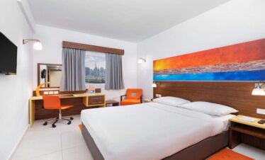 Modern double bedroom at Citymax Hotel Bur Dubai featuring a white bed with wooden headboard, orange accent chairs, a work desk, and a window view of the Dubai city skyline including the Burj Khalifa