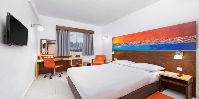 Modern double bedroom at Citymax Hotel Bur Dubai featuring a white bed with wooden headboard, orange accent chairs, a work desk, and a window view of the Dubai city skyline including the Burj Khalifa