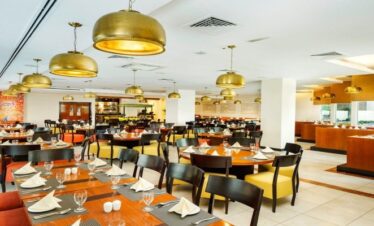 The main dining hall at Citymax Hotel Bur Dubai, showing a bright, modern interior with round wooden tables, yellow and orange chairs, and gold dome-shaped pendant lights