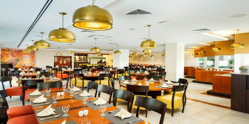 The main dining hall at Citymax Hotel Bur Dubai, showing a bright, modern interior with round wooden tables, yellow and orange chairs, and gold dome-shaped pendant lights