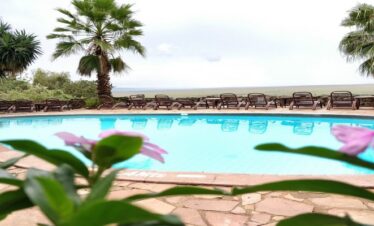 A blue swimming pool at Mara Serena Safari Lodge lined with lounge chairs, overlooking the vast plains of the Maasai Mara National Reserve under an overcast sky.