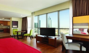 A luxury Junior Suite at Ramada by Wyndham Dubai featuring a red bed runner, a full kitchenette, a work desk, and floor-to-ceiling windows overlooking Dubai skyscrapers