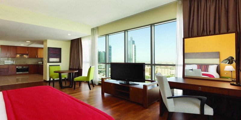 A luxury Junior Suite at Ramada by Wyndham Dubai featuring a red bed runner, a full kitchenette, a work desk, and floor-to-ceiling windows overlooking Dubai skyscrapers