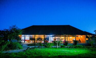 The main dining lodge of a safari camp at twilight, featuring large glass windows glowing with warm interior light, a thatched-style roof, and green grass in the foreground