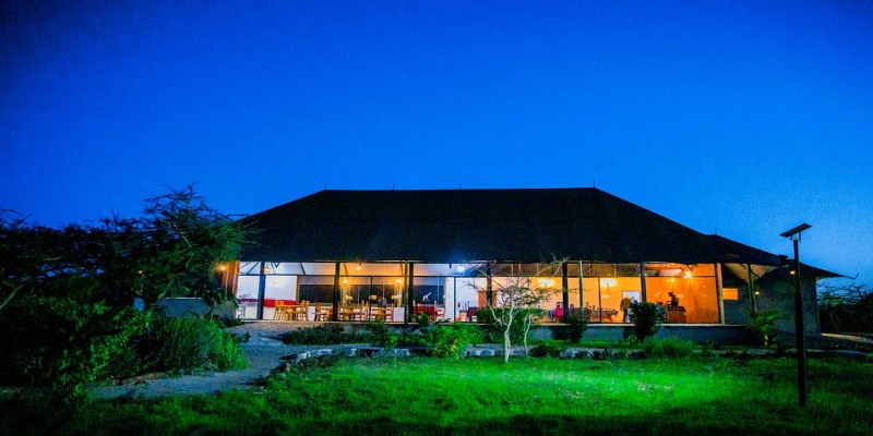 The main dining lodge of a safari camp at twilight, featuring large glass windows glowing with warm interior light, a thatched-style roof, and green grass in the foreground