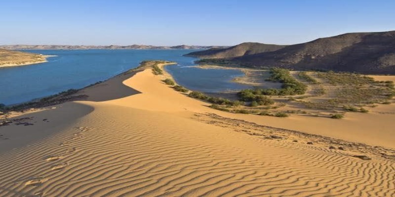 High-angle view of golden sand dunes meeting the blue waters of a desert lake in the Nyiri Desert, Kenya, with rocky hills in the background under a clear sky.